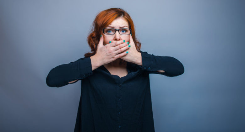 European appearance woman with glasses in a black shirt covering her mouth with her hands on a gray background, fright, fear